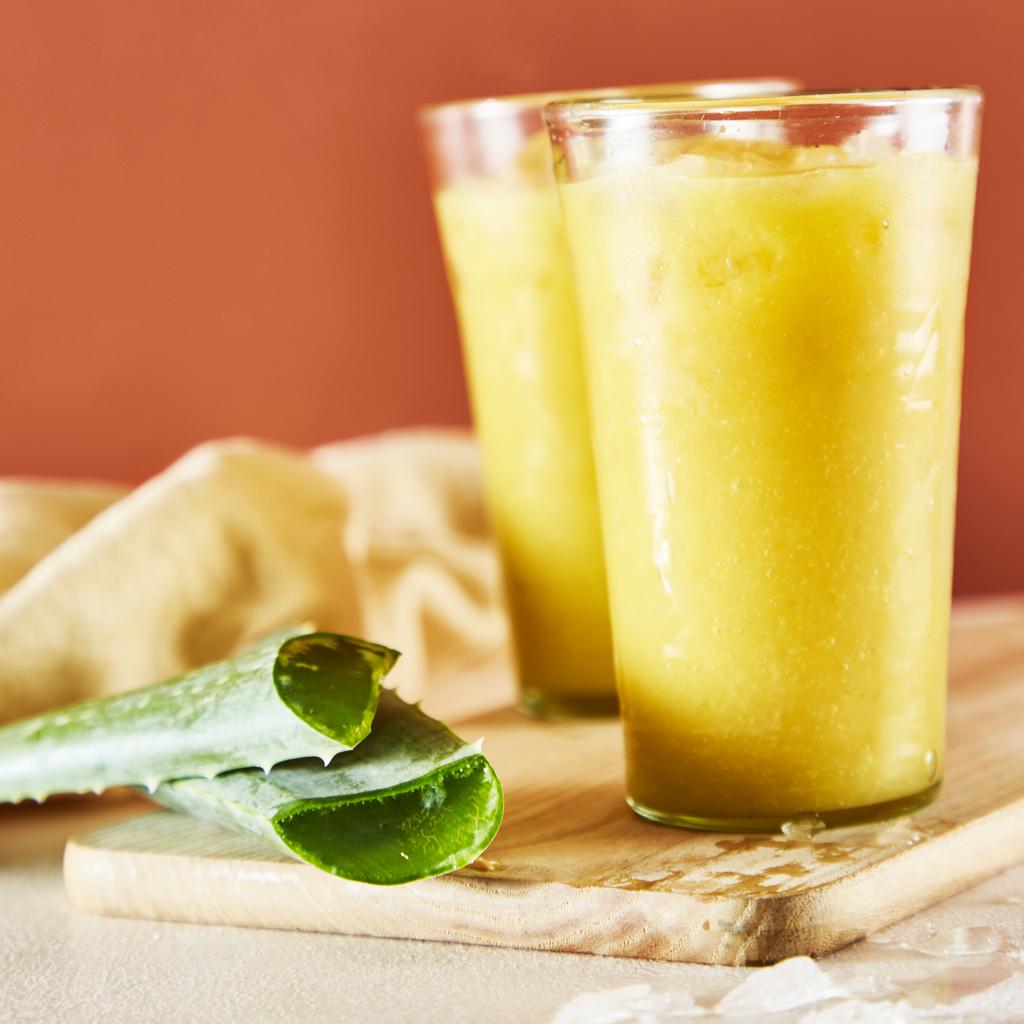 Three colorful aloe vera smoothies with tropical fruits including mango, berries, and coconut served on a beachside table.
